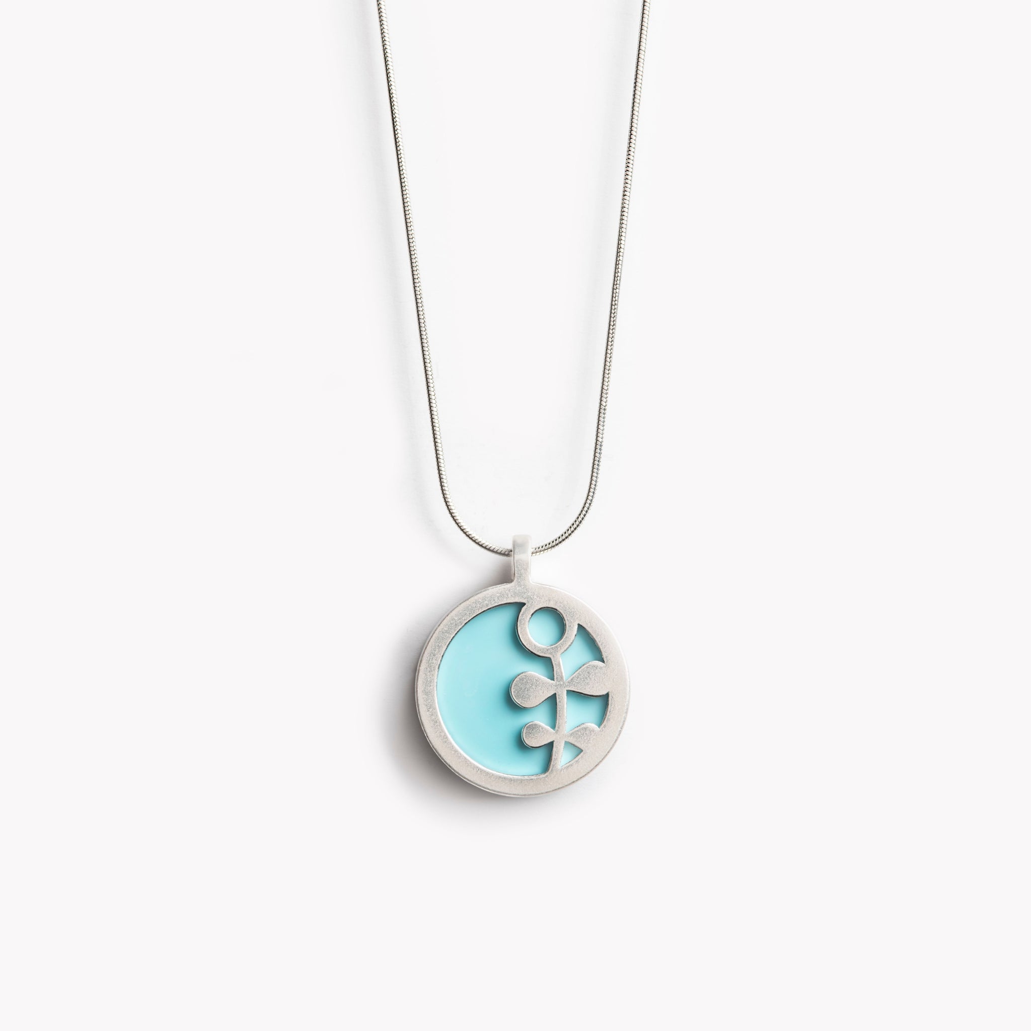 The image shows a circular pendant with a simple Scandinavian style flower stem design.  It has a pale turquoise inset which is surrounded by brightly polished pewter, with the flower  stem also highlighted in polished pewter. The pendant is bathed in sunlight on a crisp white  background. This is a simple yet vibrant, modern and colourful pendant necklace design.