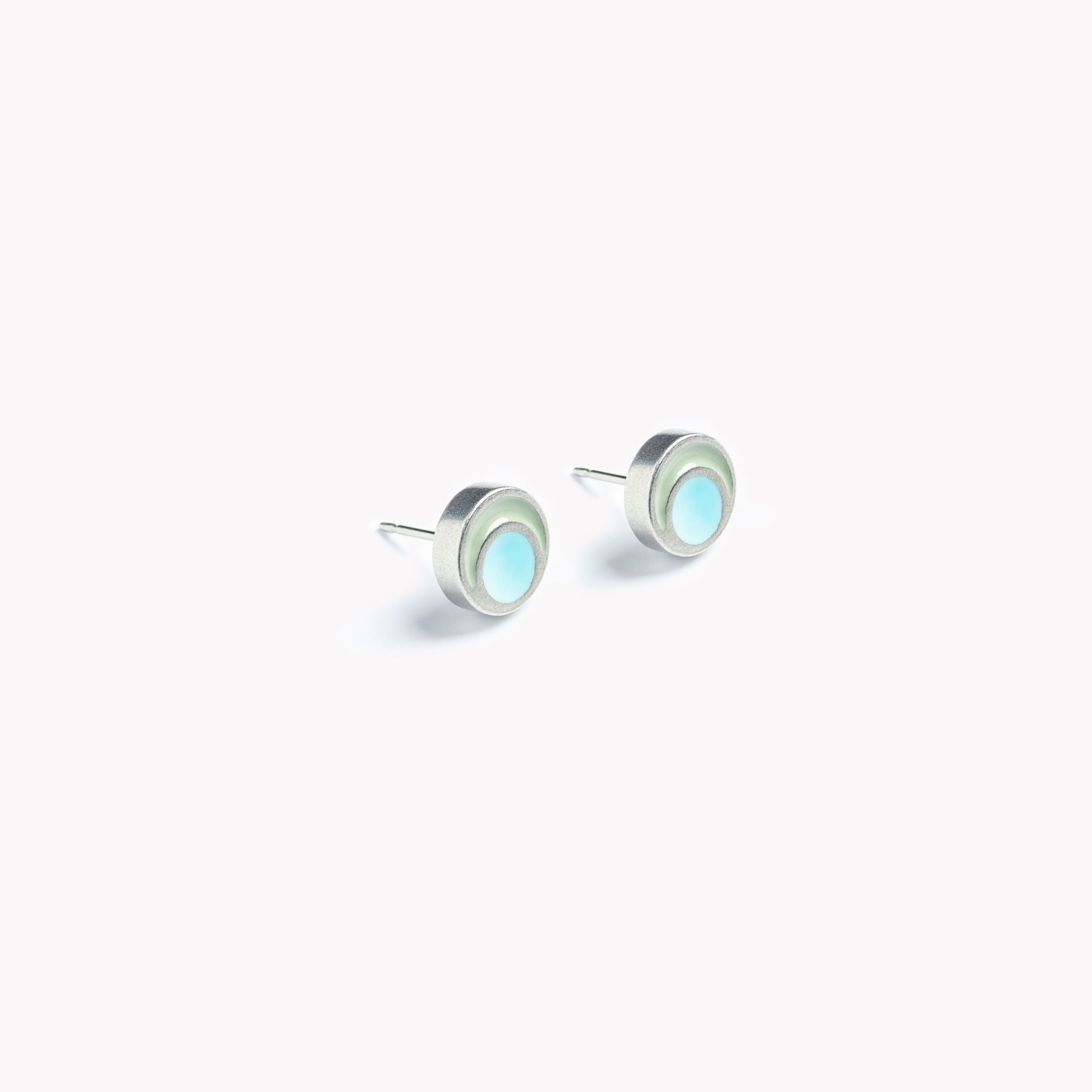 The image shows a pair of simple, vibrant, grey and turquoise circular stud earrings. Pictured  on a crisp white background, the light turquoise circular inset is surrounded by a pale grey rim  leading to a brightly polished pewter edge. The circular stud earrings are bathed in sunlight,  showing off the beautifully reflective surface of these minimal, modern and colourful  stud earrings.