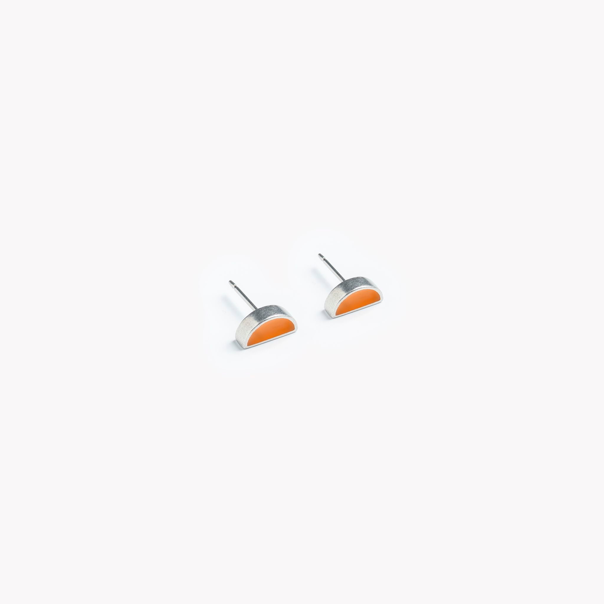The image shows a brightly coloured pair of half circle shaped orange stud earrings. The orange inset is silky smooth and runs up to a brightly polished pewter edge and surround. Pictured on a crisp white background, the stud earrings are bathed in sunlight. This is a modern, colourful, minimal stud earring design.