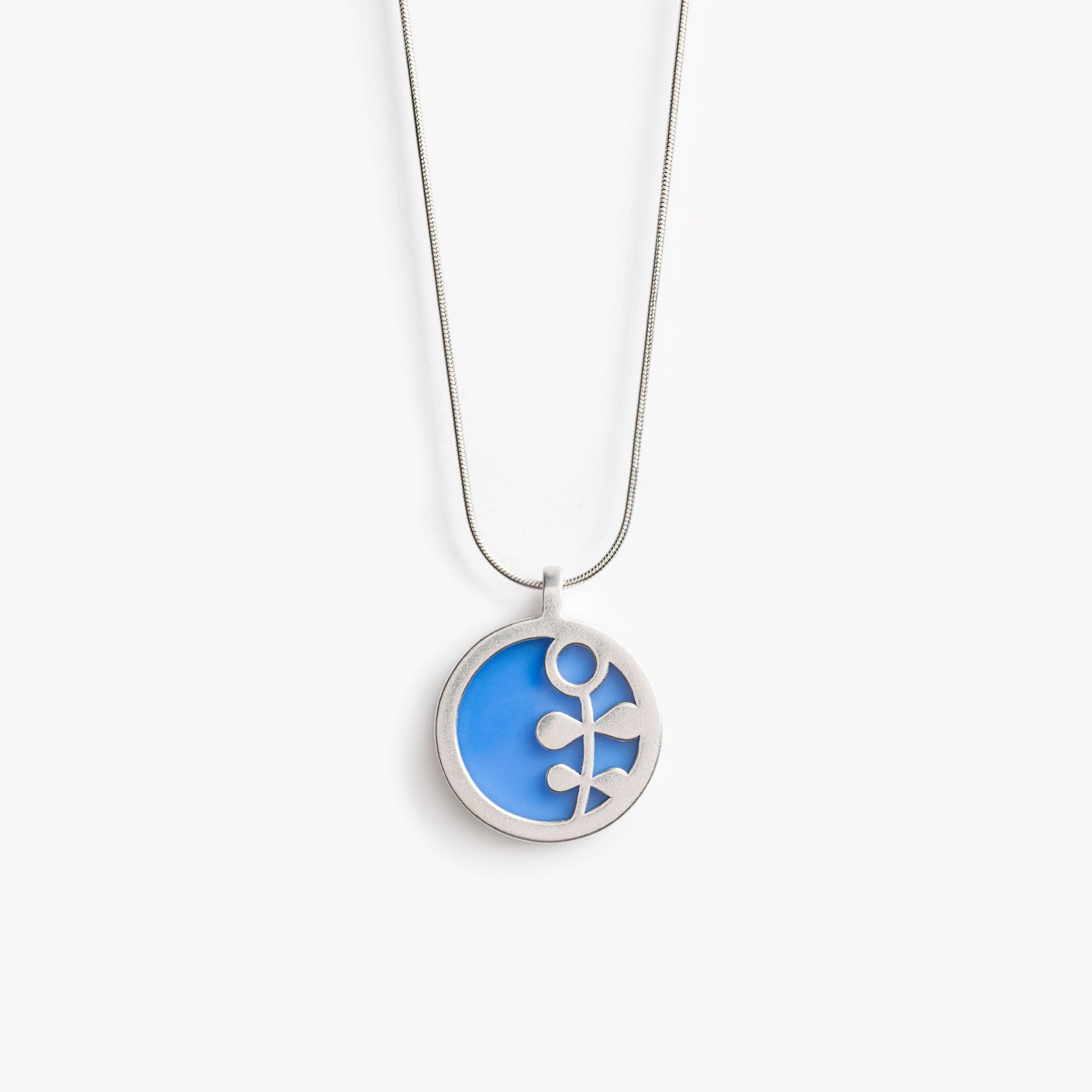 The image shows a circular pendant with a simple Scandinavian style flower stem design.  It has a coastal blue inset which is surrounded by brightly polished pewter, with the flower stem also highlighted in polished pewter. The pendant is bathed in sunlight on a crisp white  background. This is a simple yet vibrant, modern and colourful pendant necklace design.
