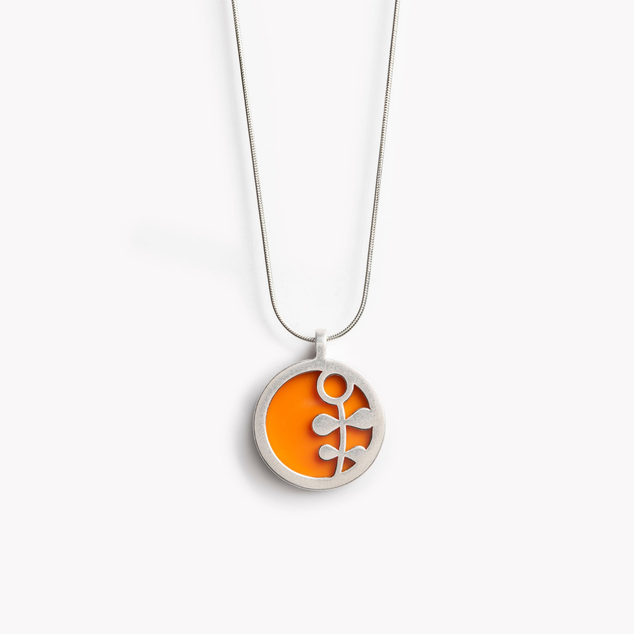 The image shows a circular pendant with a simple Scandinavian style flower stem design.  It has a citrus orange inset which is surrounded by brightly polished pewter, with the flower  stem also highlighted in polished pewter. The pendant is bathed in sunlight on a crisp white  background. This is a simple yet vibrant, modern and colourful pendant necklace design.