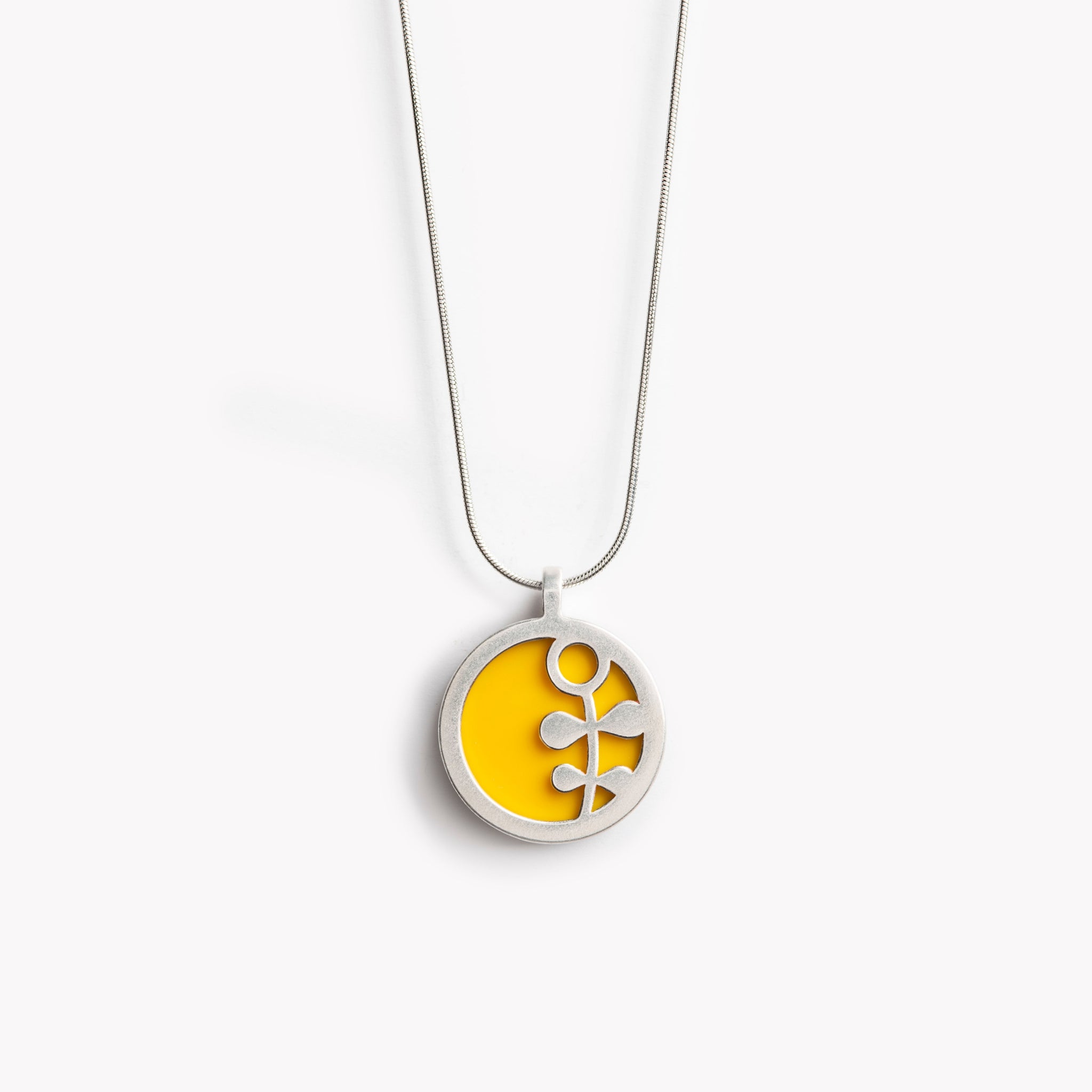 The image shows a circular pendant with a simple Scandinavian style flower stem design.  It has a bright yellow inset which is surrounded by brightly polished pewter, with the flower  stem also highlighted in polished pewter. The pendant is bathed in sunlight on a crisp white  background. This is a simple yet vibrant, modern and colourful pendant necklace design.