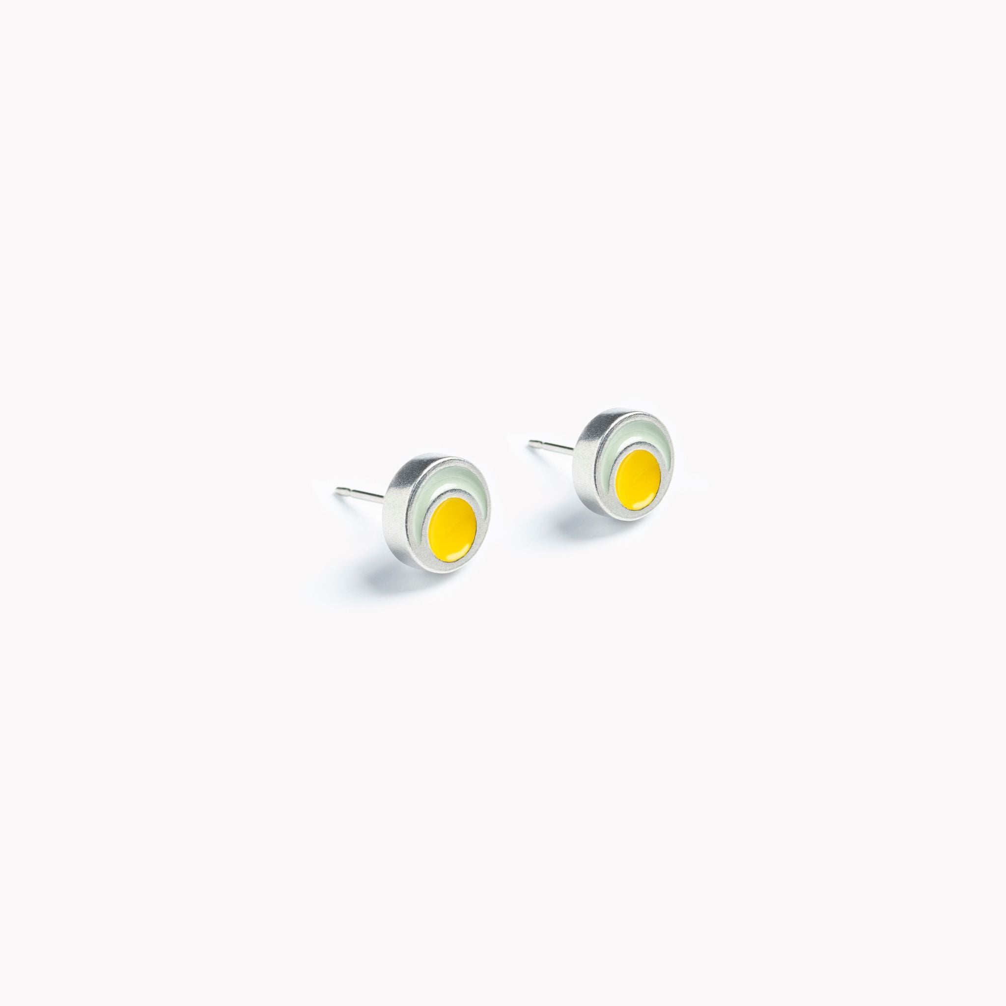 The image shows a pair of simple, vibrant, grey and yellow circular stud earrings. Pictured on a crisp white background, the bright yellow circular inset is surrounded by a pale grey rim leading to a brightly polished pewter edge. The circular stud earrings are bathed in sunlight, showing off the beautifully reflective surface of these minimal, modern and colourful stud earrings.